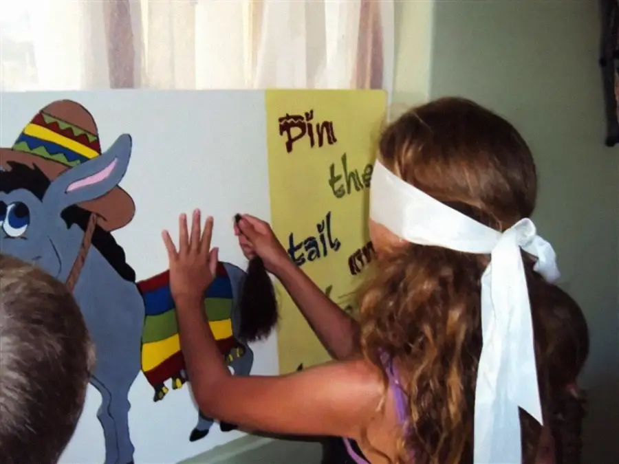 A blindfolded child plays pin the tail on the donkey at a party, reaching toward a donkey illustration with a tail in hand.