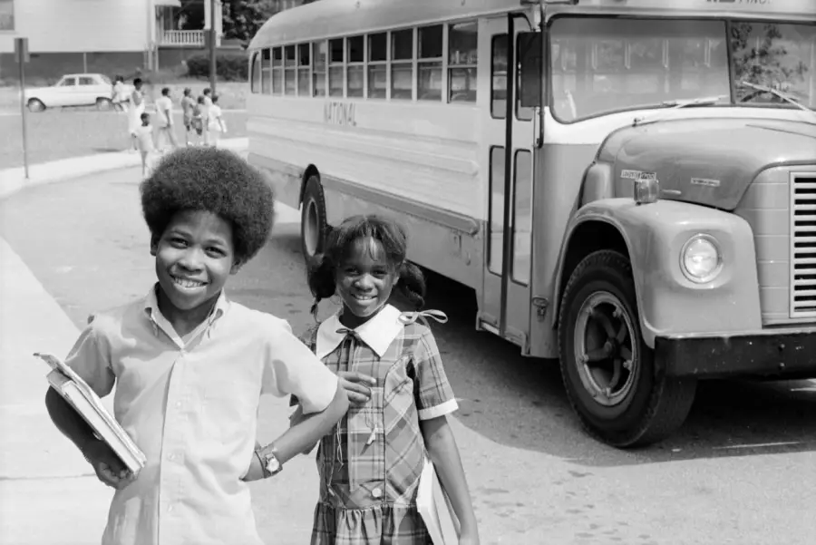 Two smiling children holding books stand in front of a school bus. More children walk on the sidewalk in the background, and houses are visible across the street. The scene appears to be from the 1970s.