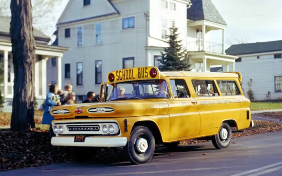 A vintage yellow school bus labeled "SCHOOL BUS" on top is driving down a suburban street with children nearby and houses in the background.