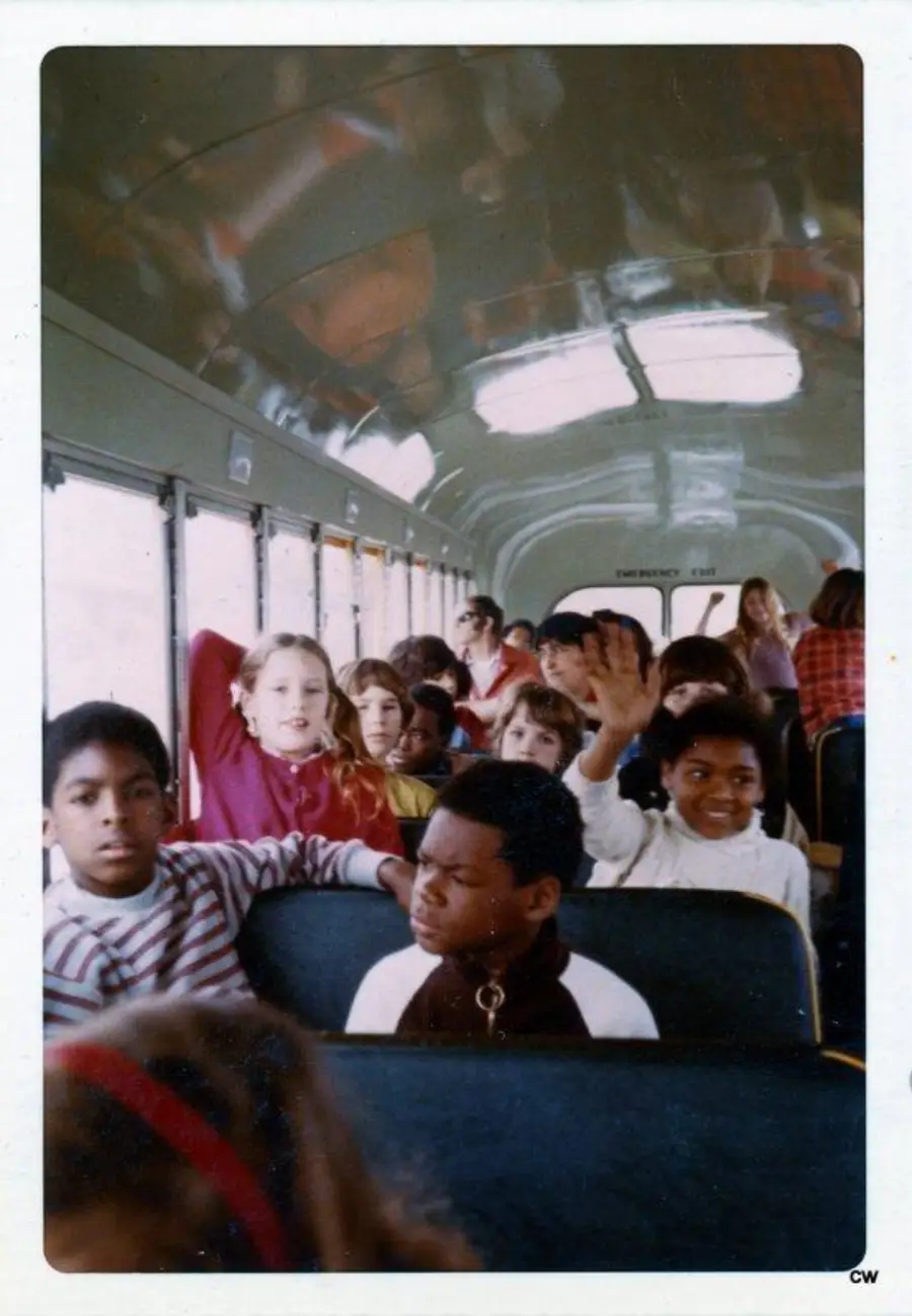 A group of children sit on a school bus. Some smile, one child waves, and others look toward the camera. The bus is mostly full, and sunlight shines in through the windows.