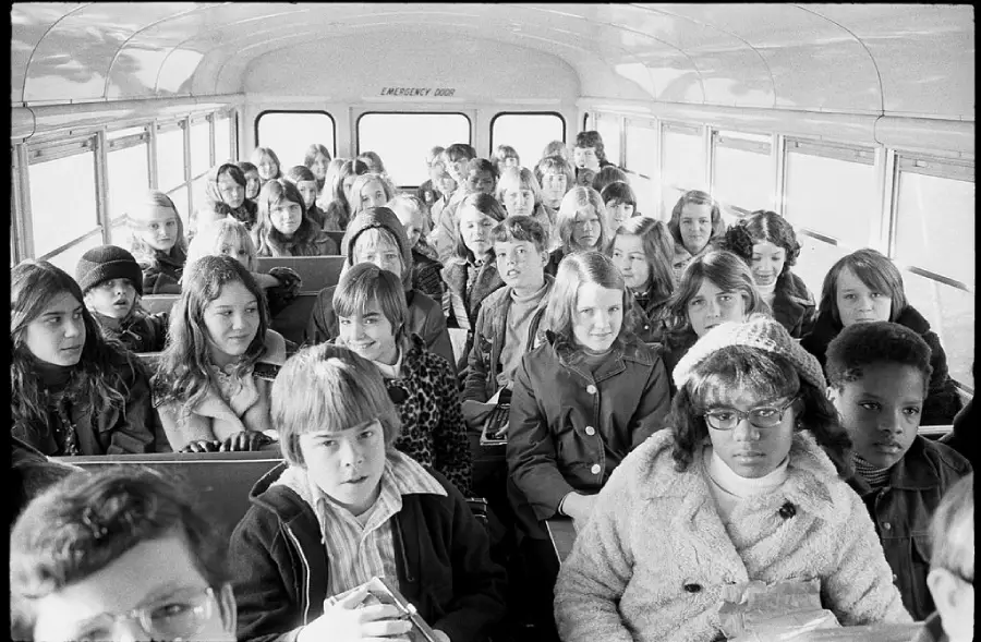 Black and white photo of a diverse group of children sitting inside a school bus, facing the camera. The students appear to be from the 1970s, wearing jackets and sweaters, with serious and neutral expressions.