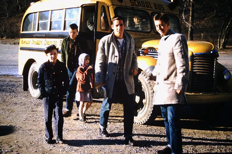 Five people, including two children and three adults or teenagers, stand outside a vintage yellow school bus on a sunny day. The group is dressed in coats, and the bus is parked on a gravel surface.