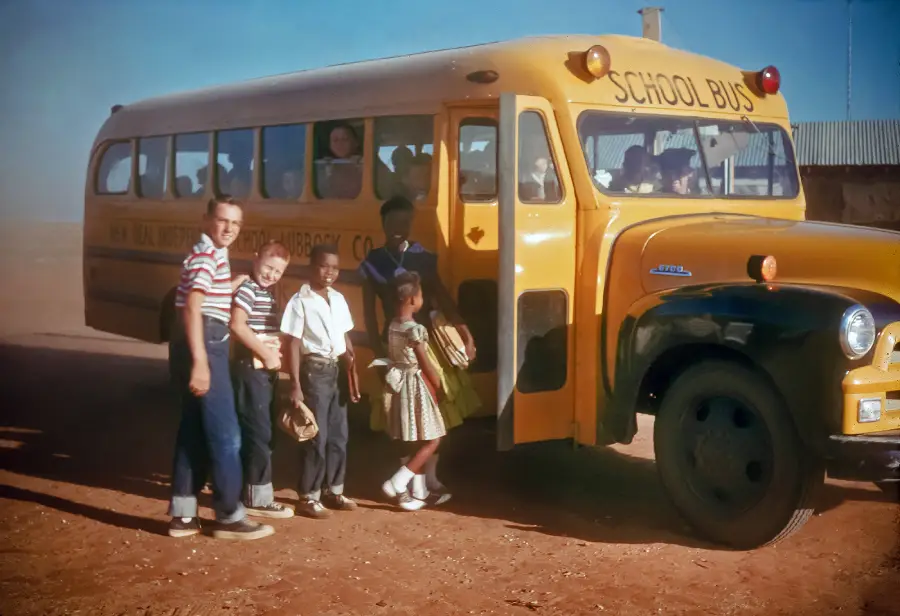 Children stand in line and board a yellow school bus on a dirt road under a clear blue sky. Some children hold paper bags, and others look out the bus windows. The scene appears to be from the mid-20th century.