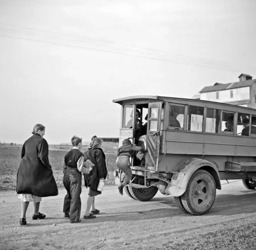 Four children walk toward and board an old-fashioned school bus on a dirt road, while an adult stands nearby. The scene is set in a rural area with a large building in the background.