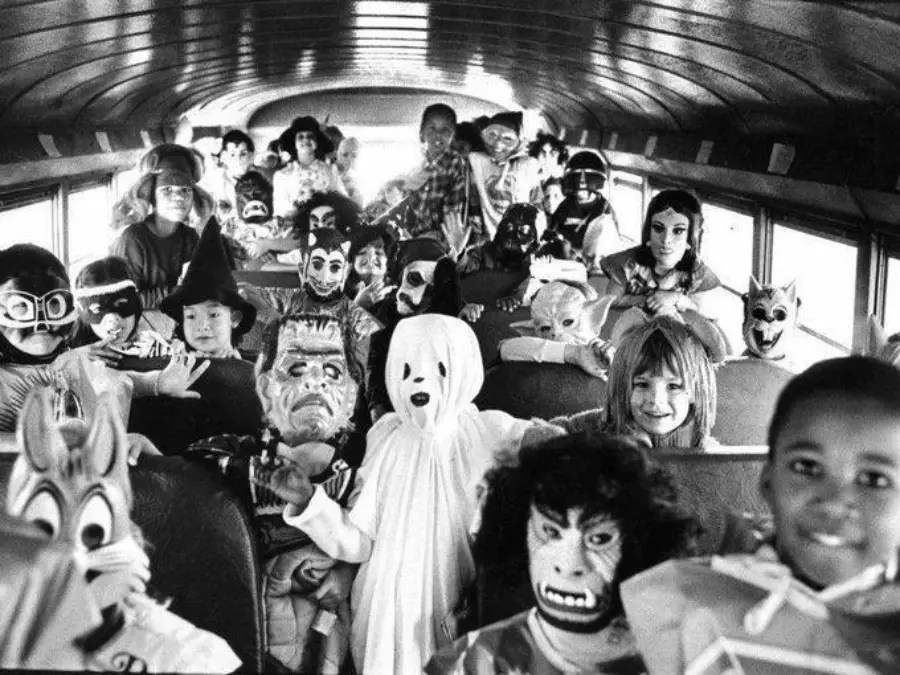 Children in various Halloween costumes and masks sit and stand inside a crowded school bus, smiling and posing for the camera. The photo is black and white, showing a festive, playful atmosphere.