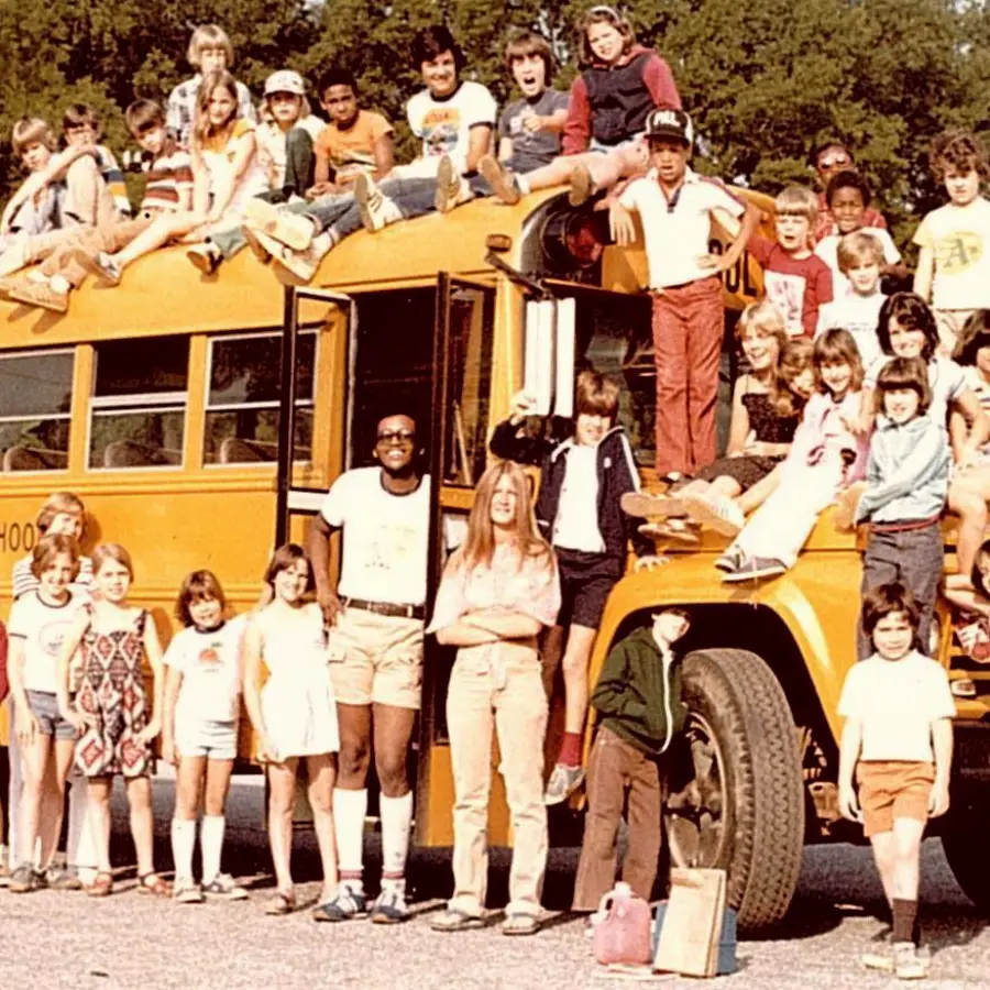 A group of children and a few adults pose on and around a yellow school bus, smiling. The photo has a vintage look, suggesting it was taken in the 1970s or 1980s. The group appears happy and relaxed.