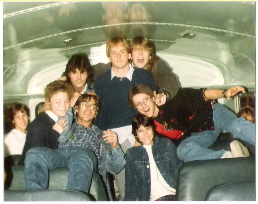A group of smiling teenagers pose together inside a school bus, some sitting and some standing, with green bus seats visible in the foreground.