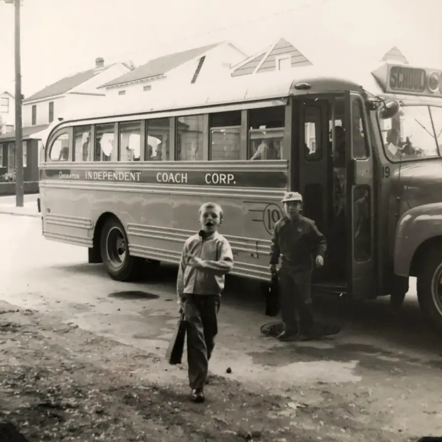 Two children in vintage clothing get off a school bus labeled “Independent Coach Corp.” One walks toward the camera, carrying a bag, while the other steps down from the bus. Houses are visible in the background.
