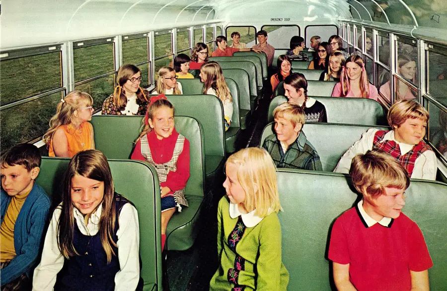 Children sit on a school bus, smiling and talking to each other. The seats are green, and the bus is brightly lit with sunlight. Most children are wearing colorful 1970s-style clothes.