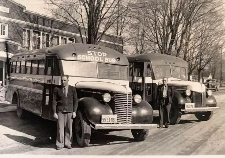 Two men stand in front of two vintage school buses parked on a tree-lined street, with buildings visible in the background. The buses have “STOP SCHOOL BUS” signs on top. It appears to be a black and white photo.