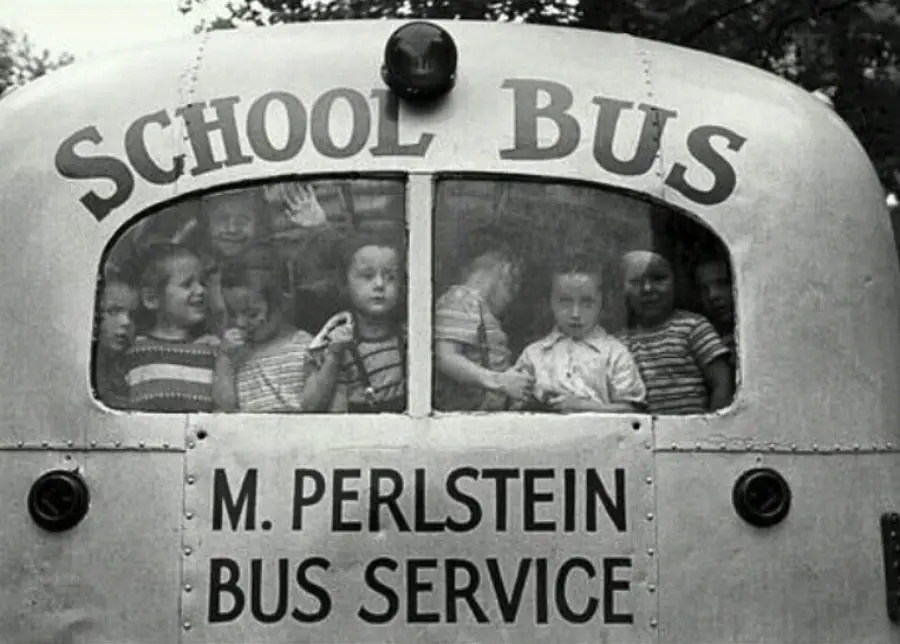 Black-and-white photo of children crowded inside a school bus, seen through the rear window. The bus has "SCHOOL BUS" and "M. Perlstein Bus Service" written on the back. Some kids look out, others smile or wave.