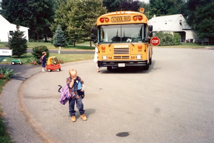 A young child with a backpack stands in front of a stopped school bus, covering their face. Two other children play nearby on a quiet neighborhood street lined with trees and houses.