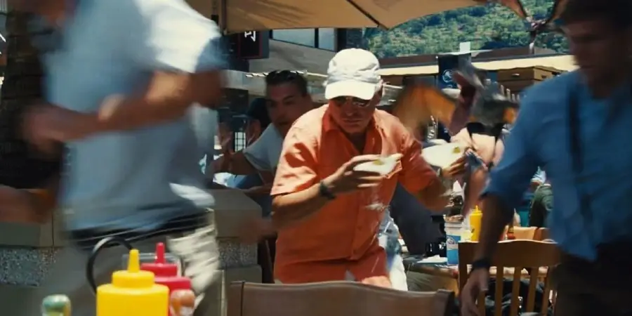 A man in an orange shirt and white hat quickly carries bowls through a busy outdoor restaurant as people rush past, with condiment bottles visible on a table in the foreground.