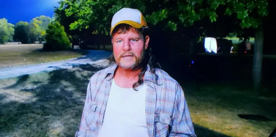 A man with a mullet haircut, wearing a white and yellow trucker cap and plaid shirt, stands outdoors on a sunny day, with trees and a dirt path in the background.