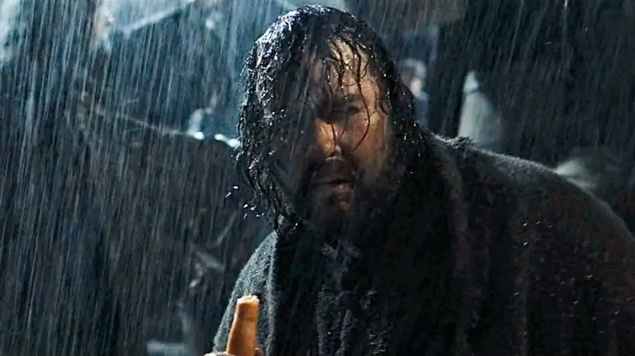 A man with wet hair and a beard stands in the rain, looking intense and holding a loaf of bread. He wears a dark, heavy coat and appears to be outdoors at night, with blurred figures and dim lighting in the background.