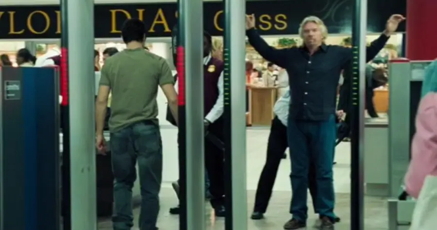 A man stands with his arms raised at an airport security checkpoint while another man walks ahead. Security staff are present, and people are visible in the background near a sign.