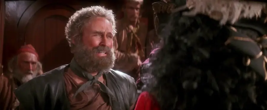 A bearded man wearing a scarf and dark vest stands indoors, looking distressed, surrounded by other people in pirate-like attire. The back of another person in a feathered hat is visible in the foreground.