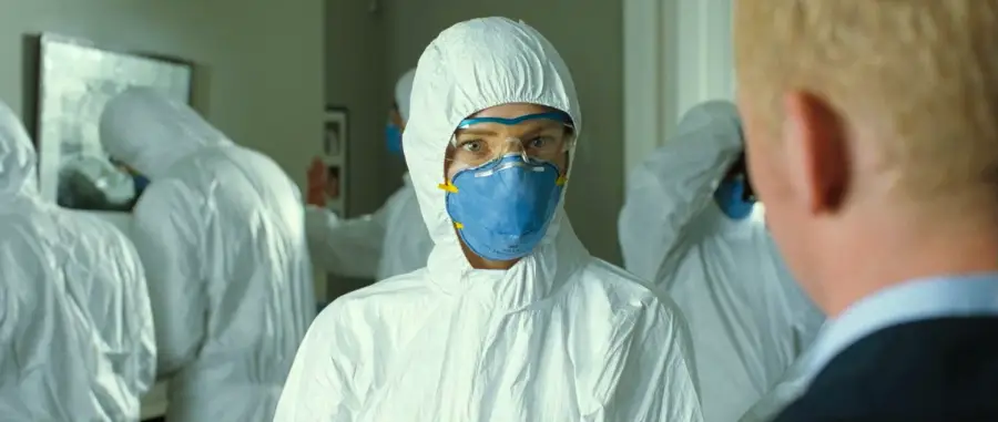 A person in a white hazmat suit, blue face mask, and goggles stands indoors facing a man in a suit. Other people in hazmat suits are visible in the background.