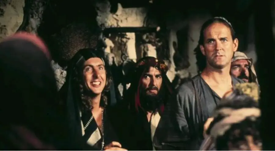 A group of people in robes stand indoors against a rough stone wall, with one man looking ahead and others gazing in different directions, some smiling and wearing head coverings.