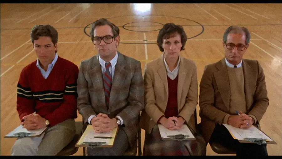 Four people sit in a row on chairs in a gymnasium, each holding clipboards and wearing formal or semi-formal attire, looking serious and attentive.