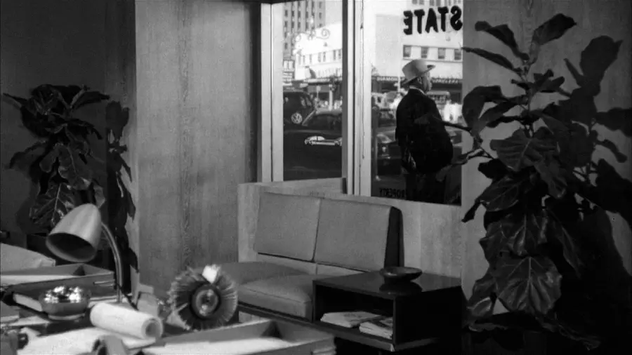A black-and-white photo of a mid-century modern office with a desk, plants, and a sofa. Through the window, a man in a hat stands outside, with buildings and cars visible on the street in the background.