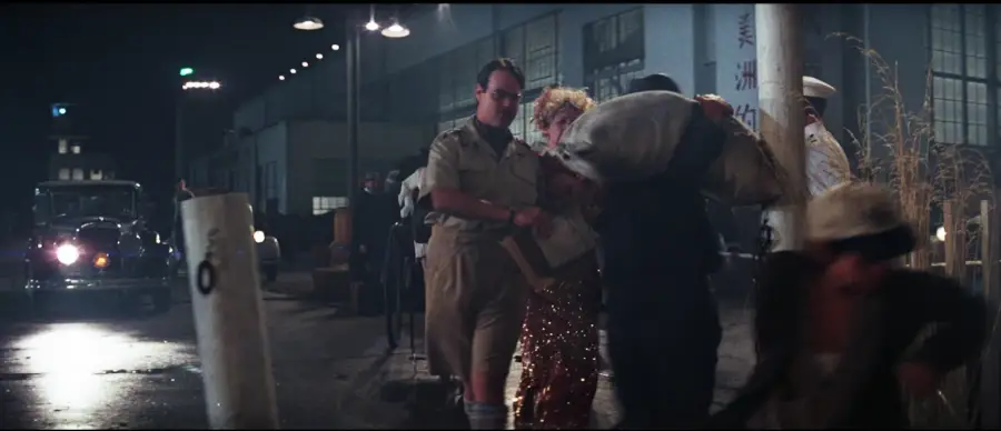 A man in a tan uniform helps a woman in a sparkling gold dress walk along a dimly lit street at night, with vintage cars and other people visible in the background.