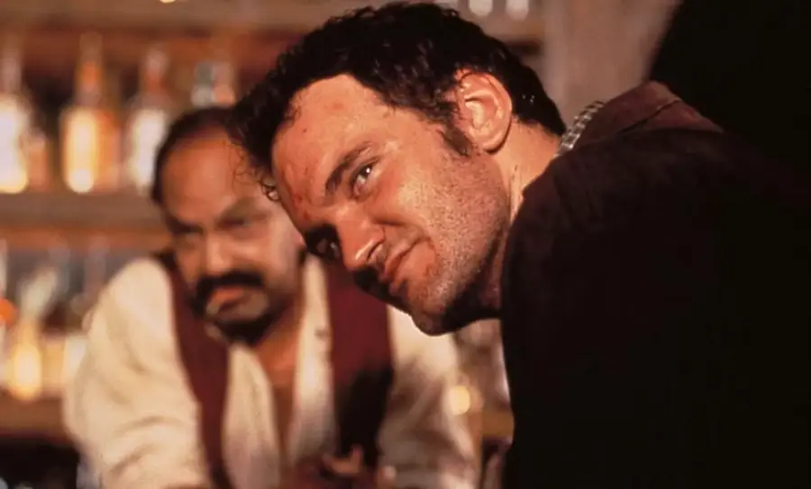 A man with a rugged appearance looks intensely to the side in a dimly lit bar, with another man in the background sitting at the bar counter. Bottles are visible on a shelf behind them.