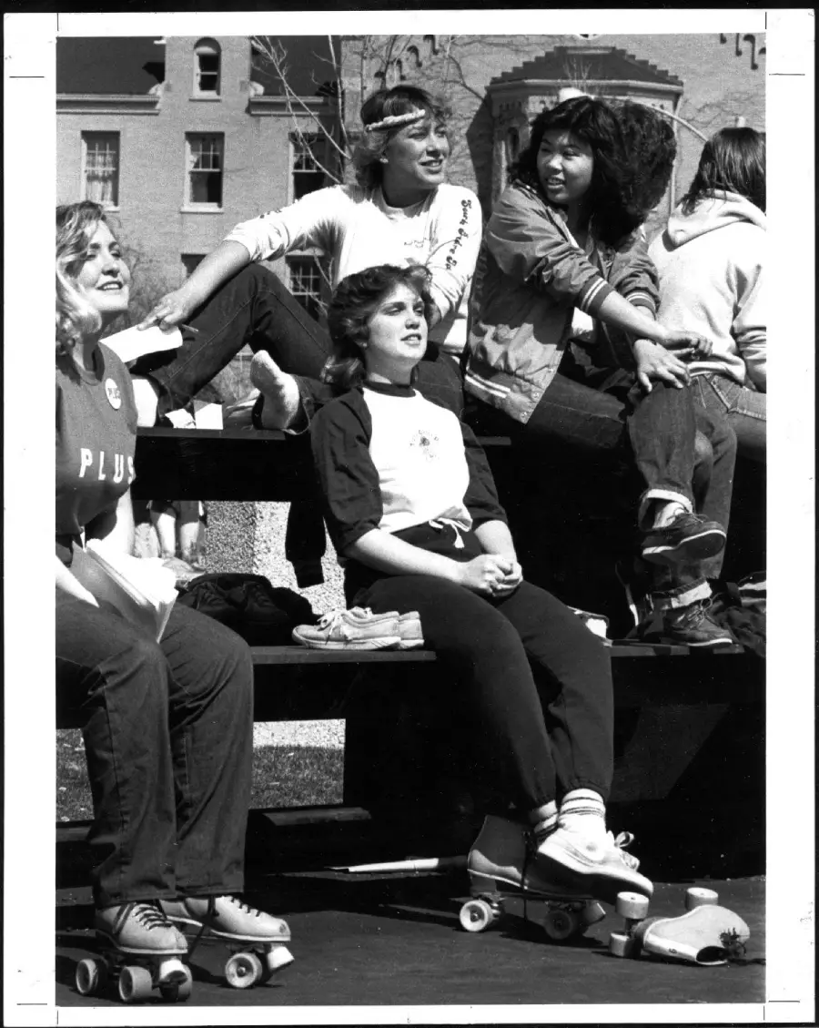 Five young people sit on bleachers outdoors, some wearing roller skates. They appear relaxed and are looking in various directions, possibly watching an event. It is a sunny day, with buildings visible in the background.
