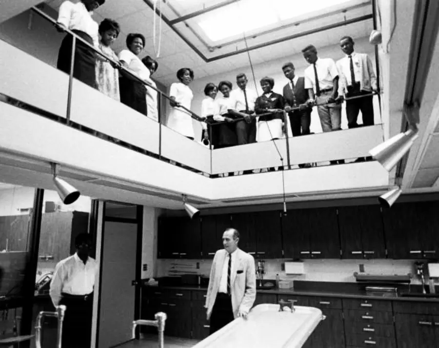 A group of people stands on an upper-level balcony observing a man in a suit below in a laboratory or medical room with a large table and overhead lights.