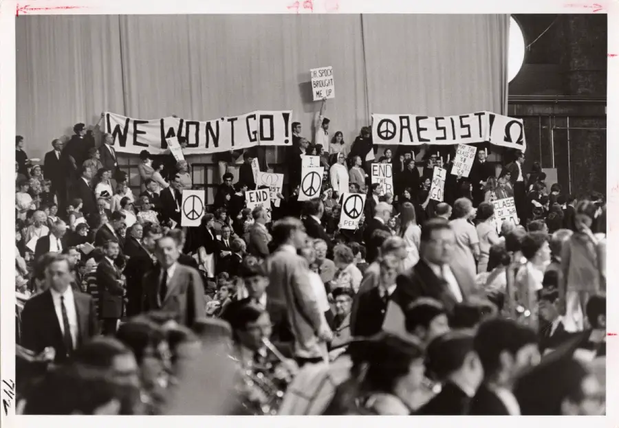 A black-and-white photo shows a crowd holding signs with peace symbols and messages like "WE WON’T GO!" and "RESIST," protesting, likely at an indoor event. Many people are seated while protesters stand.