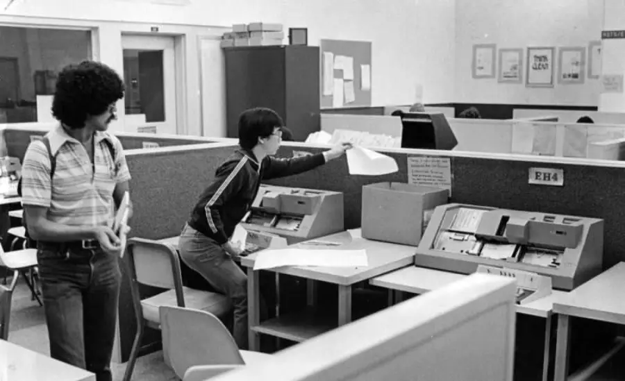 Two people in a vintage office use large computer terminals with paper printouts. One stands and looks on while the other reaches for paperwork. Cubicles and office equipment fill the room. Black-and-white photo.