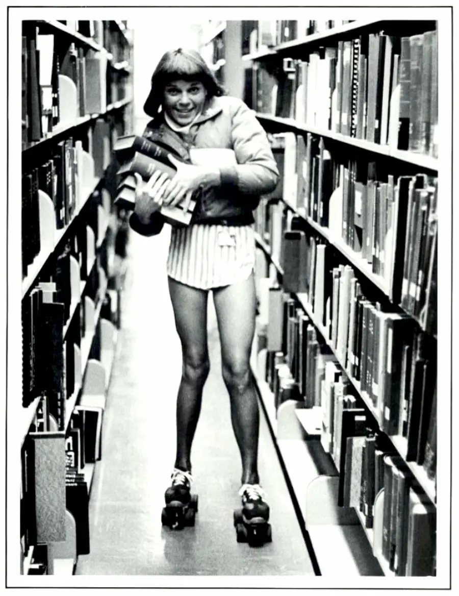 A person wearing a jacket, shorts, and roller skates stands in a library aisle, holding a stack of books and smiling. The photo is black and white.