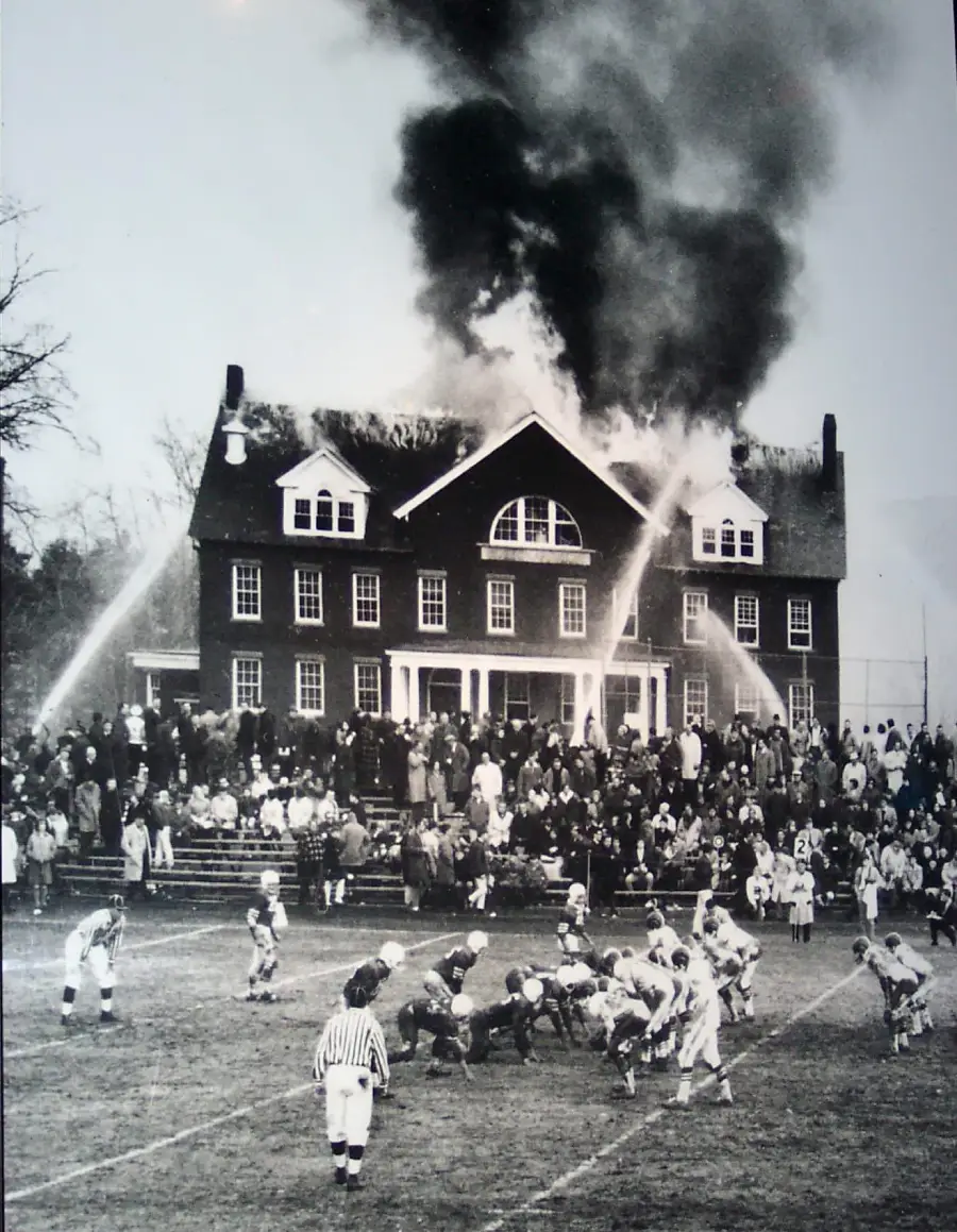 A football game is being played in front of a large building on fire, with firefighters spraying water onto the roof as a crowd watches both the game and the fire.