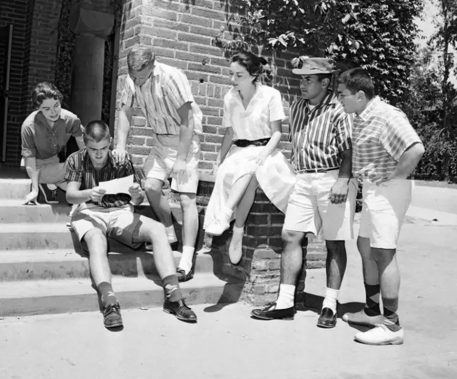 Six young adults in casual summer clothes gather on outdoor steps, looking at a piece of paper one man is holding. Some are standing, some are sitting, and they appear to be engaged and smiling.