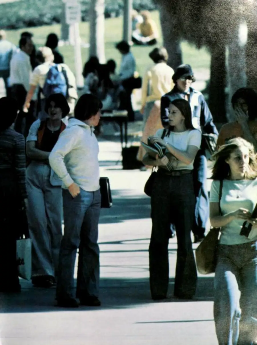 A group of people, mostly young adults, walk and talk on a sunlit outdoor path. Some carry books and bags. Trees and a lawn are in the background. The scene appears casual and busy.