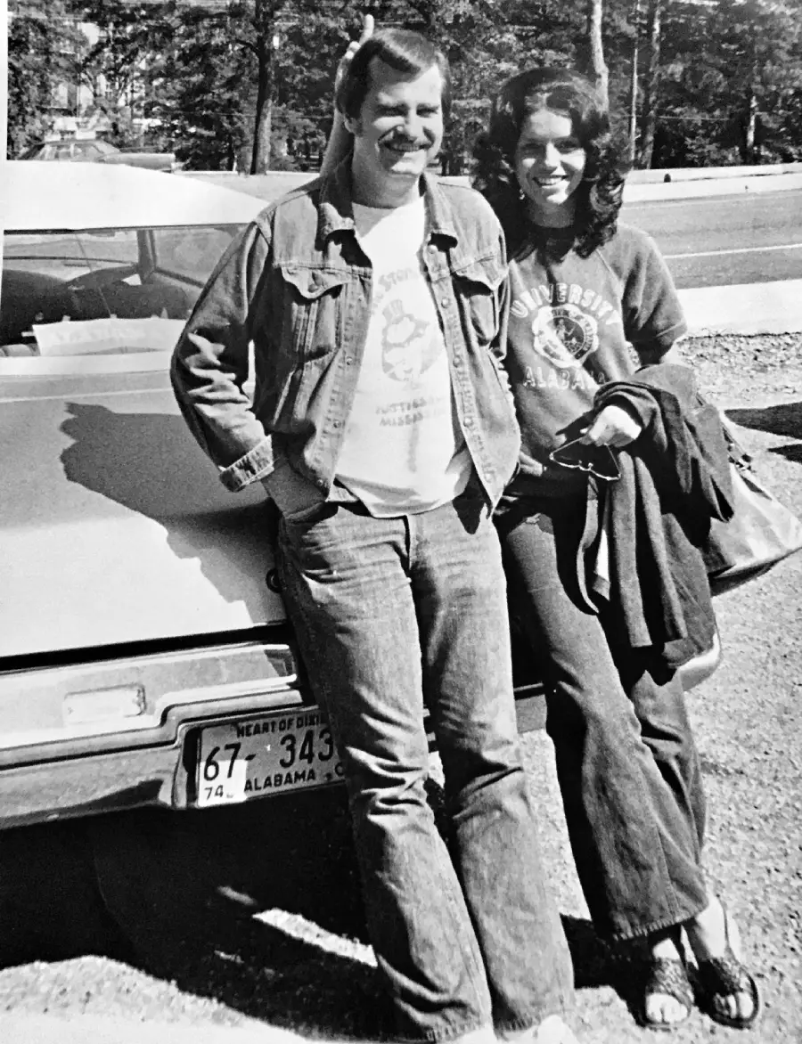 Two people in denim, one wearing a jacket and jeans, the other in a sweatshirt and holding a coat, lean against a car with Alabama plates. Trees and a road are visible in the background.