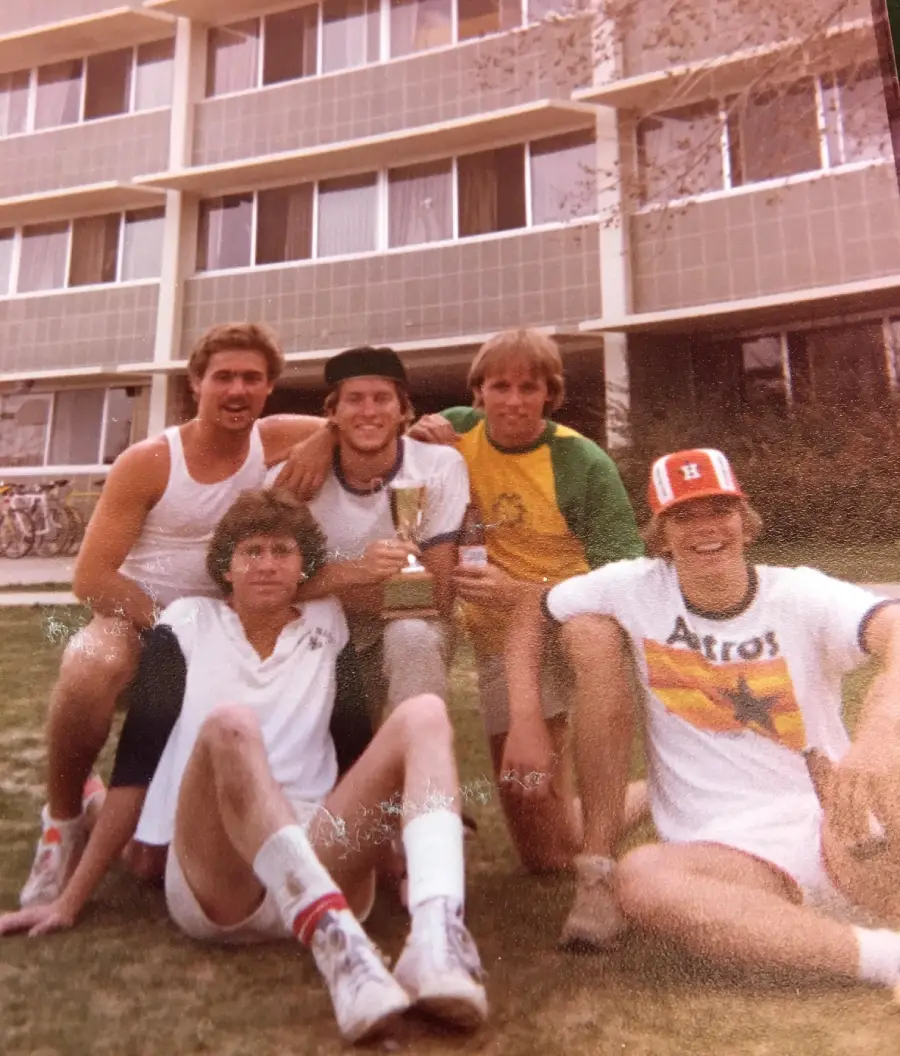 Five young men sit on grass in front of an apartment building; one holds a small trophy. They wear casual athletic clothes, and one has an Astros shirt and hat. They are smiling and relaxed.
