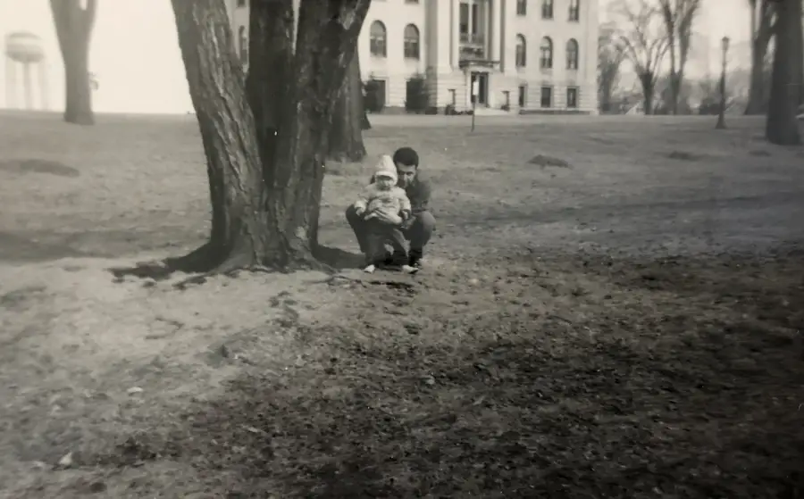 A man crouches beside a small child near a large tree in a park. An old building with columns is visible in the background. The photo is black and white.