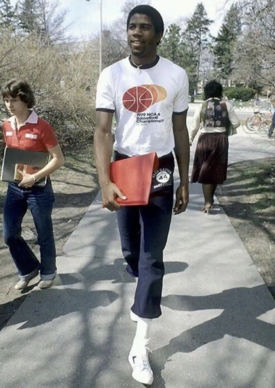 A young man in a white 1979 NCAA basketball t-shirt and blue pants walks on a campus sidewalk holding a red folder. Other students are in the background with trees and bikes visible.