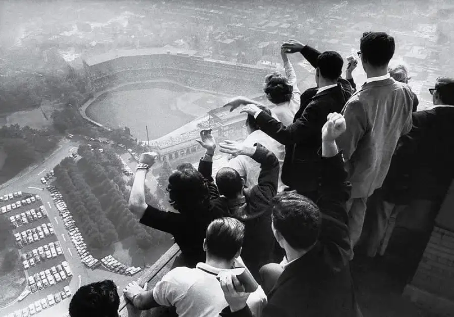A group of people stands on a high balcony, enthusiastically waving and looking down at a stadium and parking lot below on a hazy day.