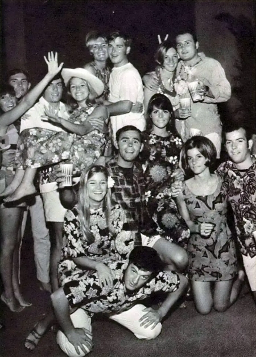 A group of young people in floral shirts pose together at a party, smiling and laughing. Some are standing, others kneeling, and a few are being carried. The photo is black and white.