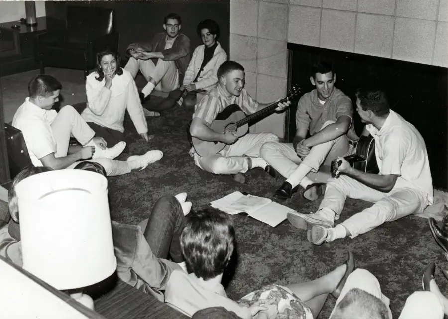 A group of young people sit in a circle on the floor, some playing guitars and others singing or listening, with papers on the carpet in a cozy indoor setting.