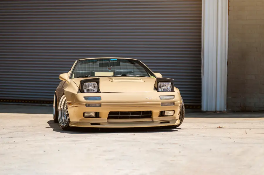 A gold Mazda RX-7 sports car with pop-up headlights is parked on a concrete surface in front of a large, closed, gray industrial garage door.