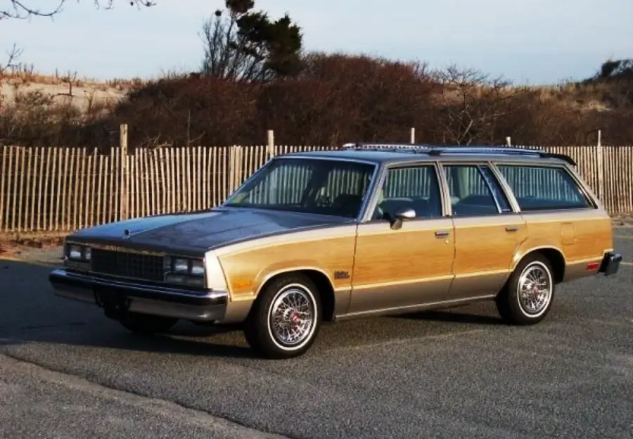 A vintage station wagon with wood paneling on the sides is parked on a paved road near a wooden fence and some trees.