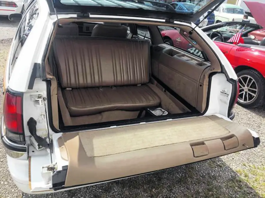 The image shows the open rear hatch of a station wagon with a rear-facing third-row bench seat in brown vinyl upholstery. The seat backs onto the open trunk area, and the car is parked near other vehicles.
