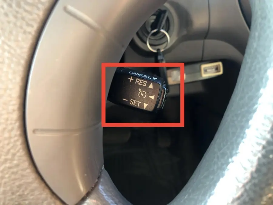 Close-up of a car’s cruise control lever behind the steering wheel, showing options to cancel, resume, and set speed. A key is inserted in the ignition.