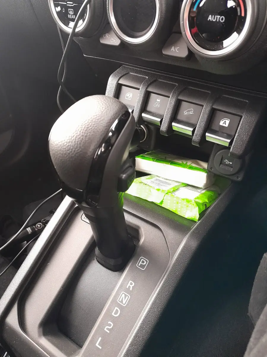 A car’s gear shift lever is in the center console near the AC controls. A green pack of tissues is stored in a compartment under four toggle switches.