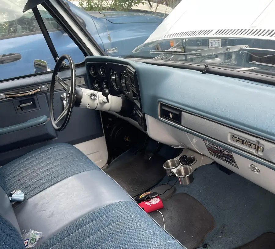 Interior of a vintage pickup truck with blue upholstery, a large steering wheel, dashboard controls, cup holders, and keys on the seat. The hood is up, and another blue vehicle is visible outside the window.