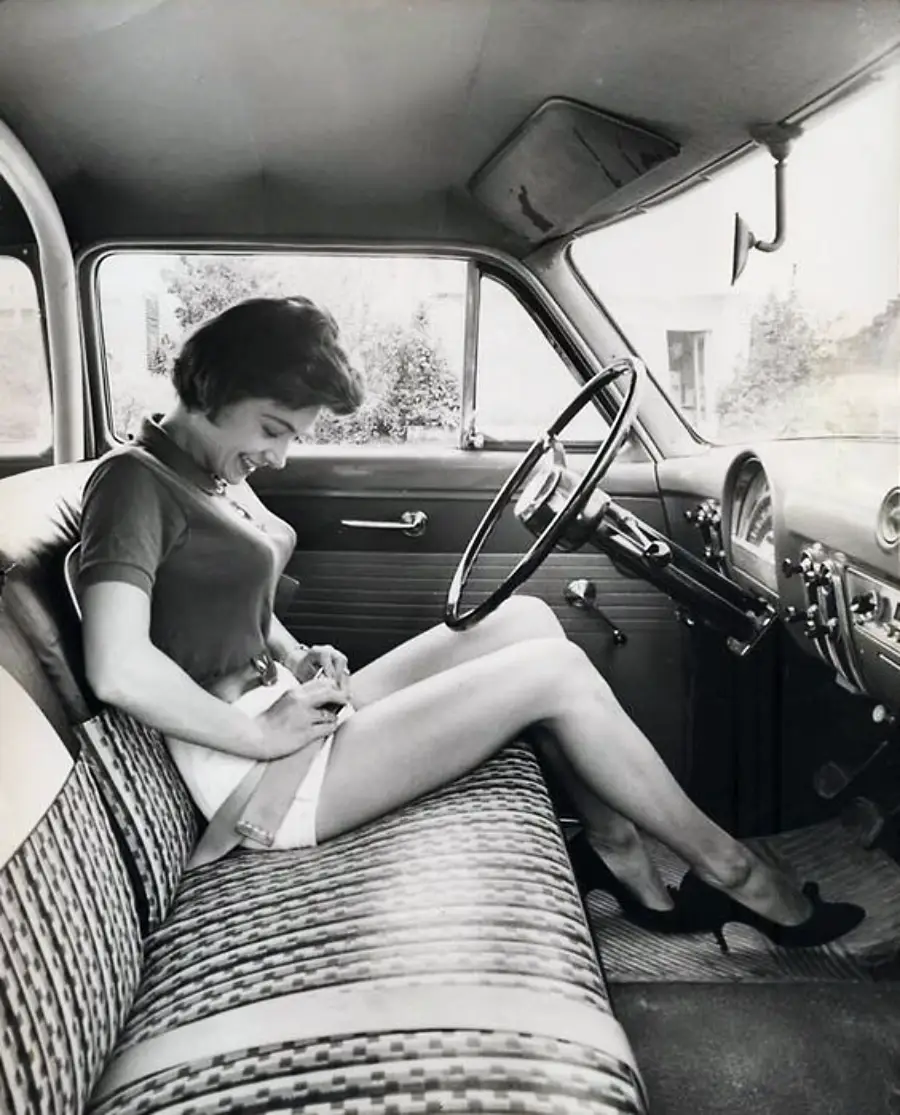 A woman in a 1950s car sits on a patterned bench seat, fastening her seatbelt. She wears a blouse, shorts, and heels, and is smiling while looking down at the belt.