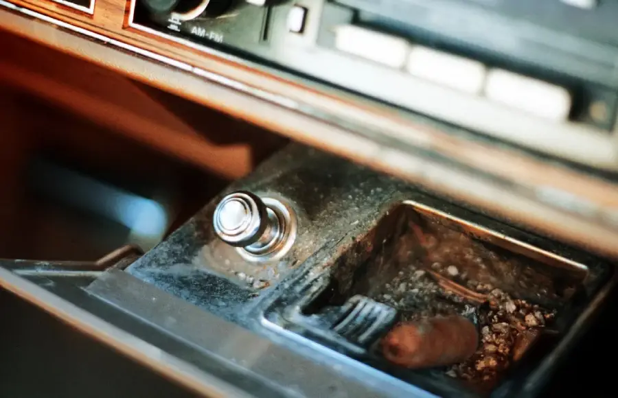 A close-up of a car’s old, dirty ashtray filled with ashes and a cigarette butt, next to a metallic car cigarette lighter in the dashboard.